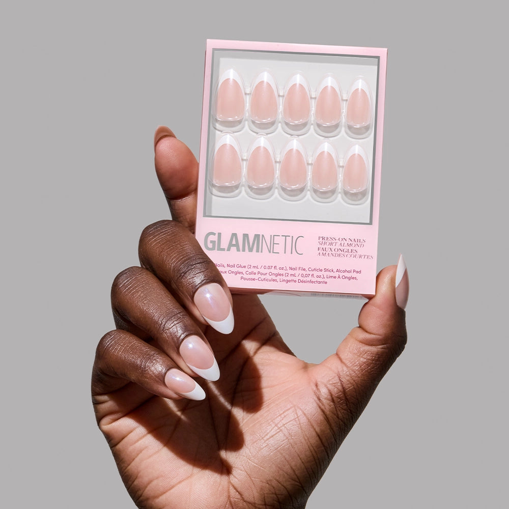 A hand with long, almond-shaped French tip nails holds a glamnetic "Mrs." press-on nail set featuring ten pink French-tip nails—an ideal chic bridal look—against a plain grey background.